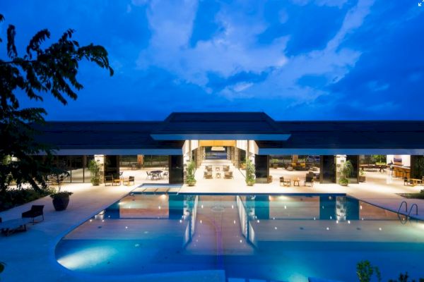 A luxurious, modern building with an illuminated pool area at dusk, surrounded by seating and greenery, under a dramatic sky.