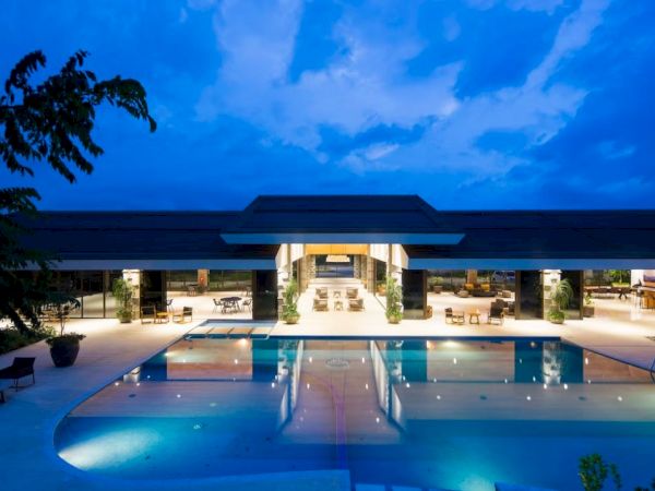 A luxurious, modern building with an illuminated pool area at dusk, surrounded by seating and greenery, under a dramatic sky.