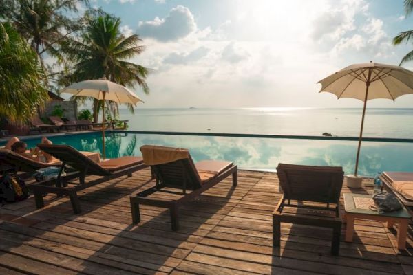 Poolside chairs and umbrellas on a wooden deck next to an infinity pool, overlooking a serene ocean view with palm trees and a bright sky.