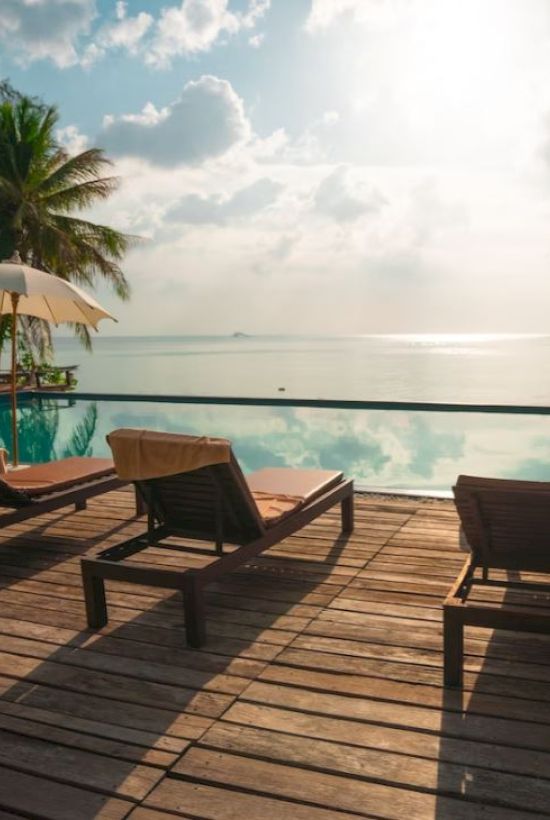 The image shows lounge chairs and umbrellas by an infinity pool overlooking a serene ocean view, with palm trees and a partly cloudy sky.