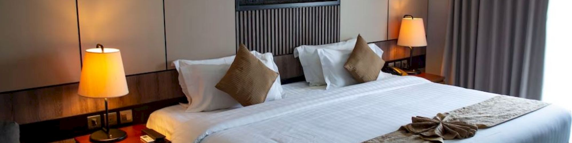 A hotel room featuring a neatly made bed with brown accents, bedside lamps, and a decorative wall piece above the headboard ends the scene.
