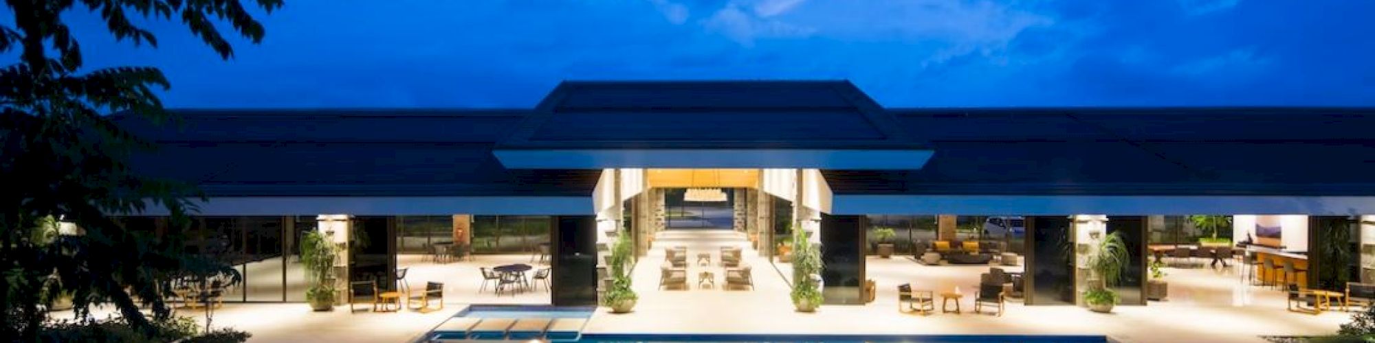 An illuminated, modern building with large, open spaces and a spacious swimming pool in front, set against a twilight sky.