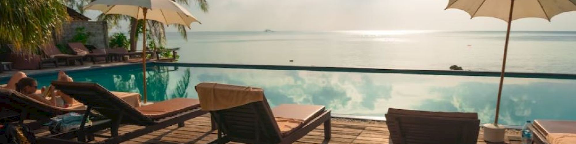 A tranquil beach scene with lounge chairs and umbrellas by a pool, overlooking a serene ocean with a clear sky and scattered clouds.