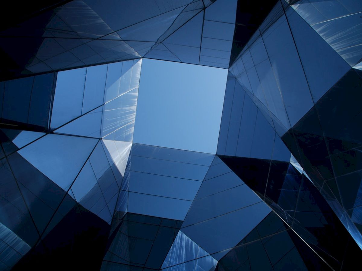 The image shows a geometric architectural structure with reflective surfaces, creating a view of the sky through the complex, angular design.