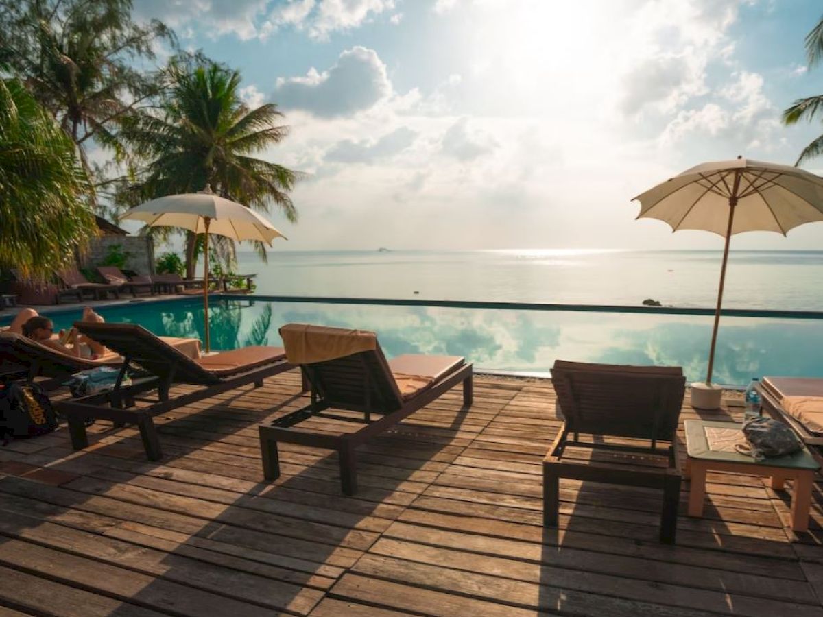 A serene poolside setup with lounge chairs, umbrellas, and a stunning view of the sky and water, surrounded by palm trees.