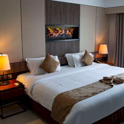 A hotel room with a large bed, two bedside tables with lamps, pillows, a runner, and wall art above the bed. There is a curtain on the right.