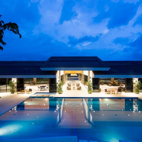 A modern building with a large, illuminated pool in the foreground and an open, airy design under an evening sky.