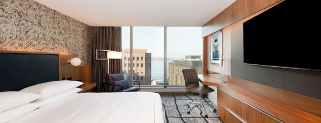 A modern hotel room with a neatly made bed, large TV, work desk, and floor-to-ceiling windows offering a city view.
