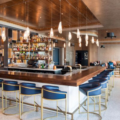 This image shows a modern bar with blue bar stools, a well-stocked liquor shelf, hanging lights, and a lounge area with additional seating.
