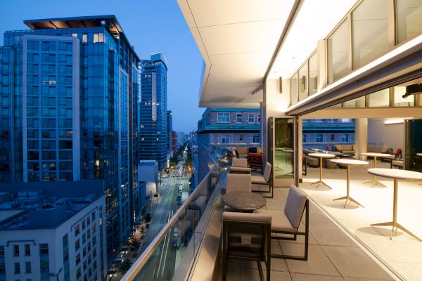 A modern urban balcony at dusk, with seating areas and tables, overlooking a city street lined with high-rise buildings.