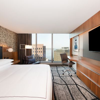 A modern hotel room featuring a large bed, a wall-mounted TV, a desk with a chair, and a large window with a view of city buildings.