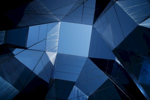 This image showcases an upward view of a modern, glass-covered building structure, creating a geometric pattern against a clear blue sky.