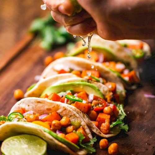 The image shows several tacos filled with chickpeas, diced vegetables, and garnished with avocado slices, as a hand squeezes lime juice on them.