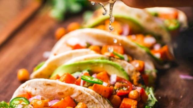 The image shows several tacos filled with chickpeas, diced vegetables, and garnished with avocado slices, as a hand squeezes lime juice on them.
