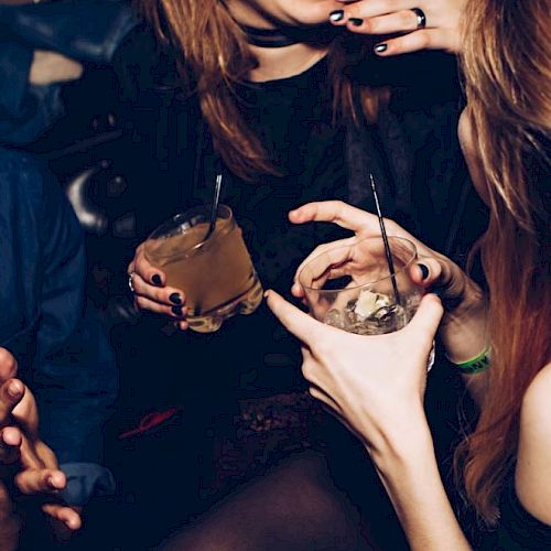 People are gathered, holding drinks and socializing in a lively setting, possibly a party or bar. They appear to be enjoying their time together.