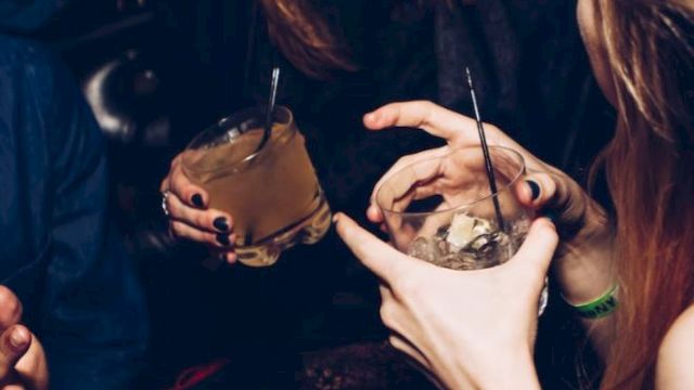 People are gathered, holding drinks and socializing in a lively setting, possibly a party or bar. They appear to be enjoying their time together.
