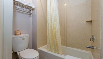 A small bathroom with a bathtub, shower curtain, toilet, and wall-mounted shelving holding towels. There is also a towel on the bathtub.