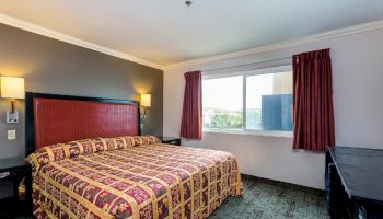 A hotel room with a king-sized bed, nightstands, lamps, a large window with red curtains, and a dresser.