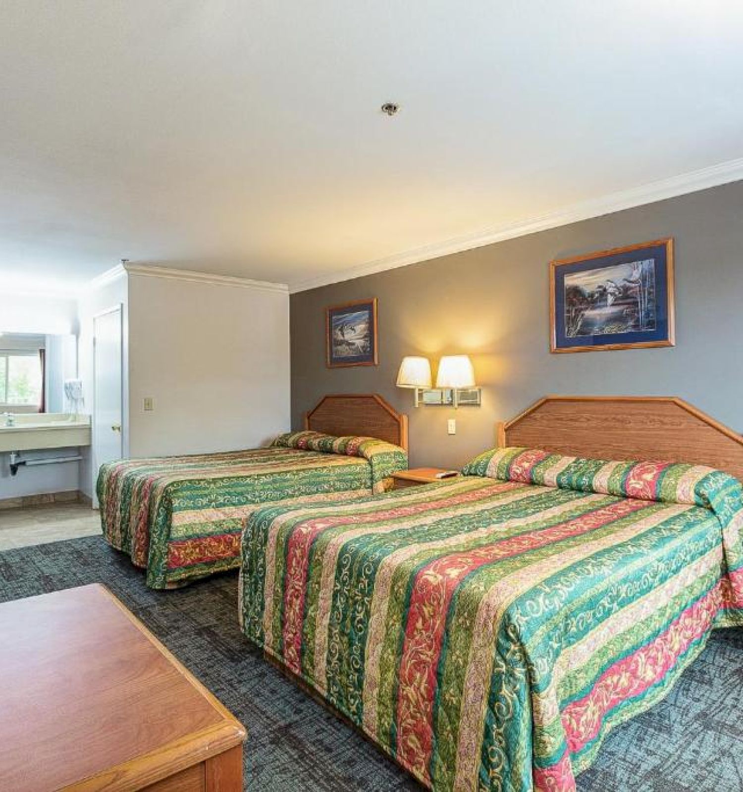 A hotel room with two beds, colorful bedspreads, wall pictures, a dresser, a TV, an armchair, and a sink area visible in the background.