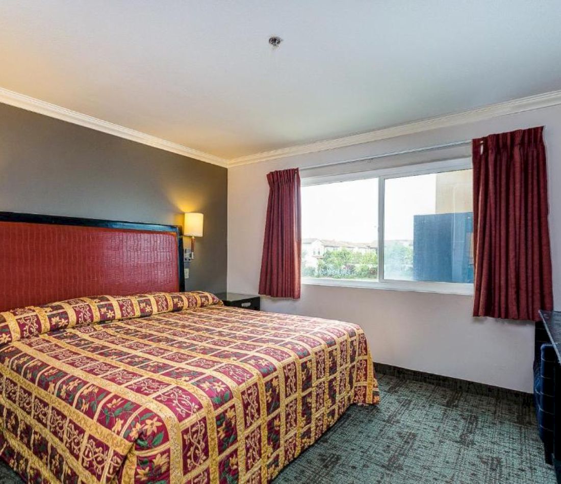 A hotel room with a large bed, a window with red curtains, and a lamp on the wall.