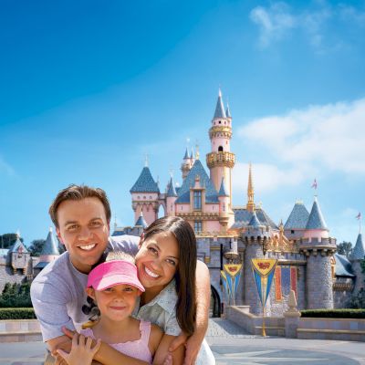 A smiling family stands in front of a castle at a theme park, enjoying a sunny day together.