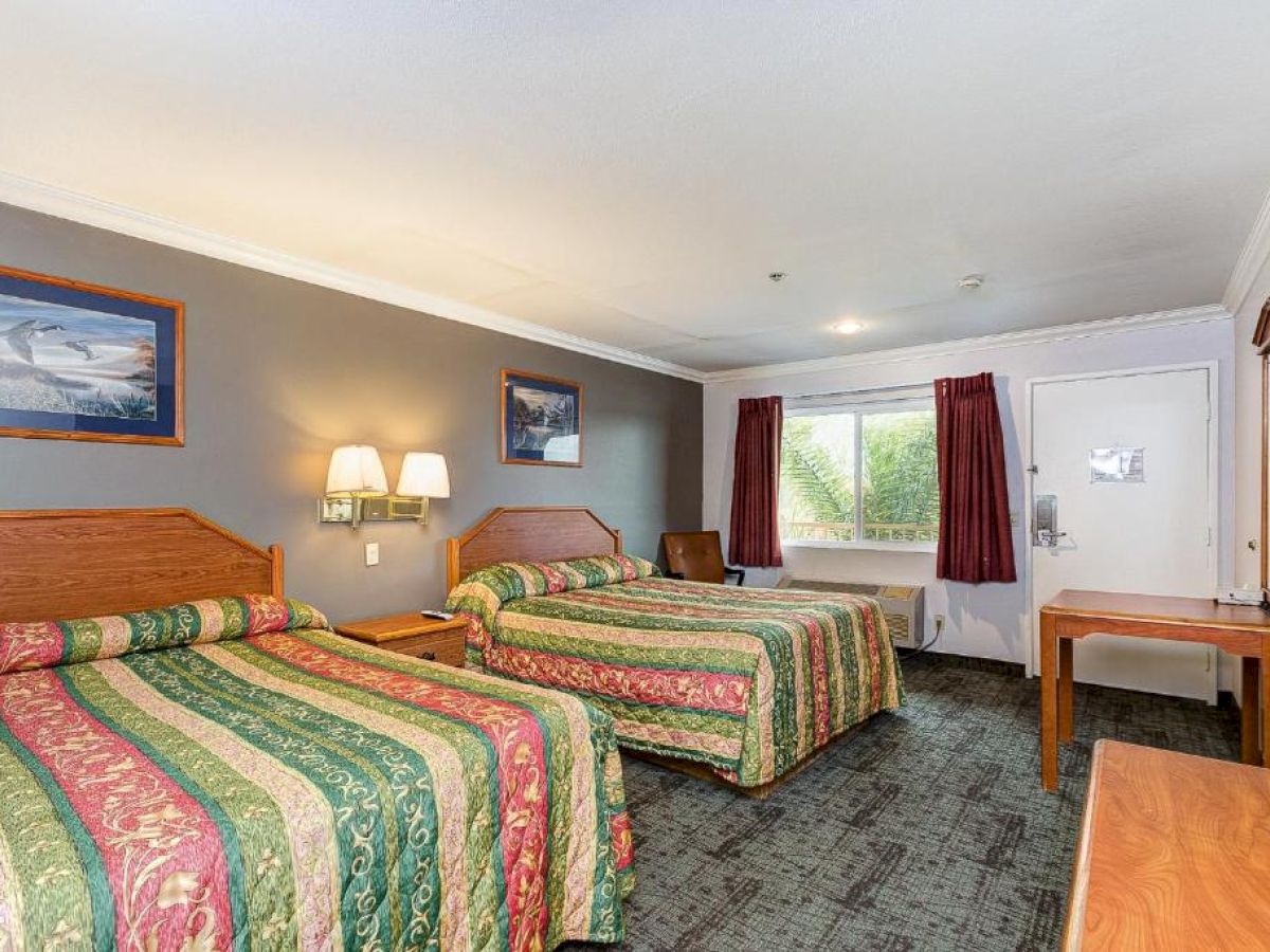 A hotel room with two beds with colorful bedspreads, wall-mounted lamps, a window, a table, a chair, and framed art on the wall.