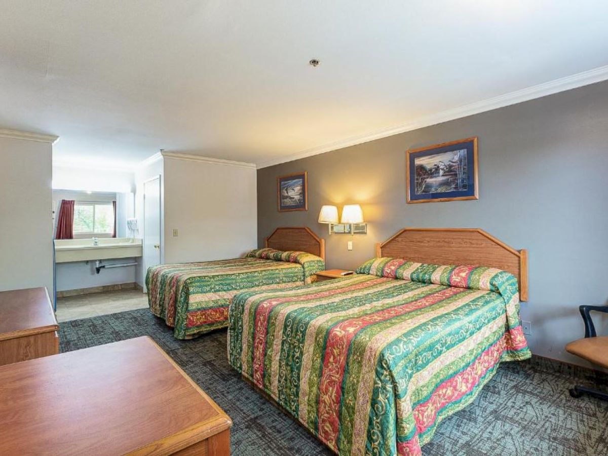 A hotel room with two double beds, colorful bedspread, a TV, a chair, a sink area, and two wall paintings illuminated by a lamp.