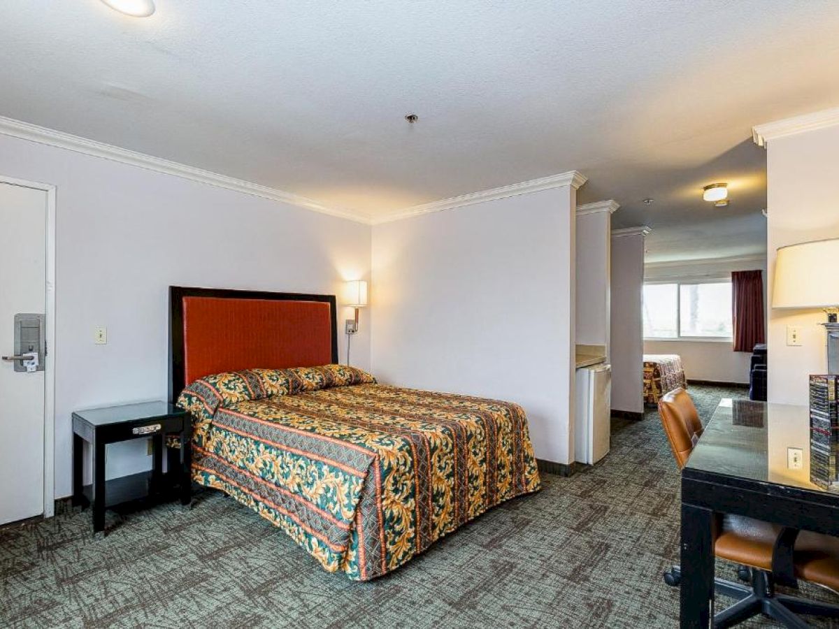 The image shows a hotel room with a bed, lamp, desk, chair, and an adjoining area leading to another room and window, creating a spacious setting.