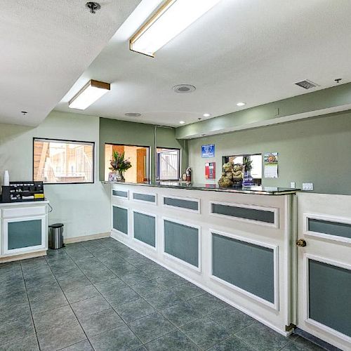 This image shows a hotel reception area with a tiled floor, a front desk, and a coffee station on the side. There is seating in the background.