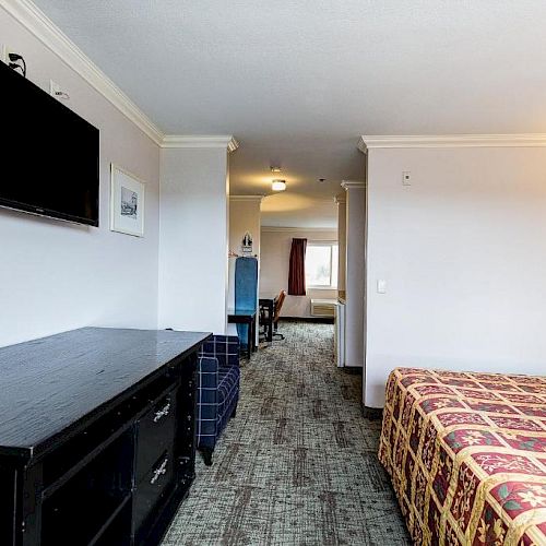 A hotel room with a bed, a wall-mounted TV, a dresser, and a view of a seating area and window in the background.