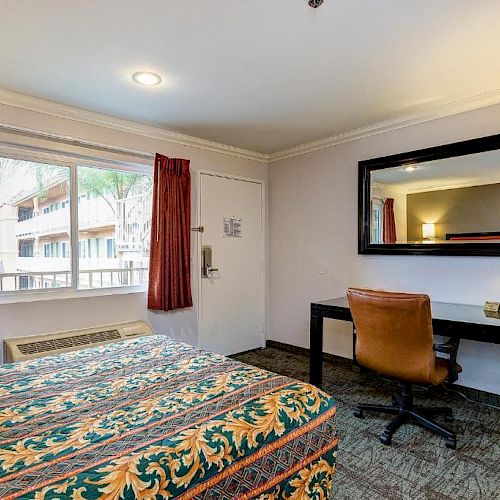 A hotel room with a bed, desk, chair, mirror, lamp, and a window with red curtains. There are books on the desk and an air conditioner visible.