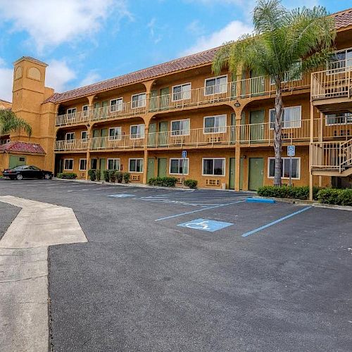 An exterior view of a multi-story building with a staircase, palm trees, and an empty parking lot under a partly cloudy sky.