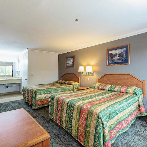 A hotel room with two beds, a TV, two lamps, framed pictures, a sink, a chair, and a table. The beds have colorful bedspreads.