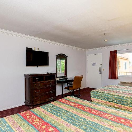 A hotel room with two beds, a TV, a desk with a chair, a window with curtains, and a drawer set is visible in this image.