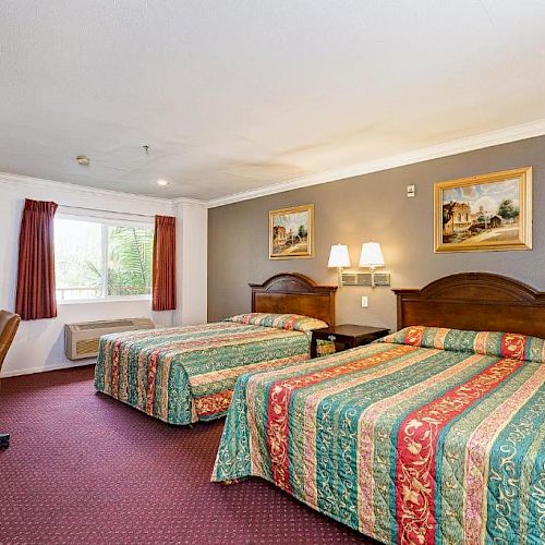 A hotel room with two double beds, colorful bedding, a desk, a chair, a dresser, lamps, and framed artwork on the wall, with natural light.