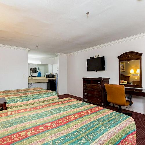 The image shows a hotel room with two beds, a TV on a dresser, a desk with a chair, and a view of a kitchenette in the background.