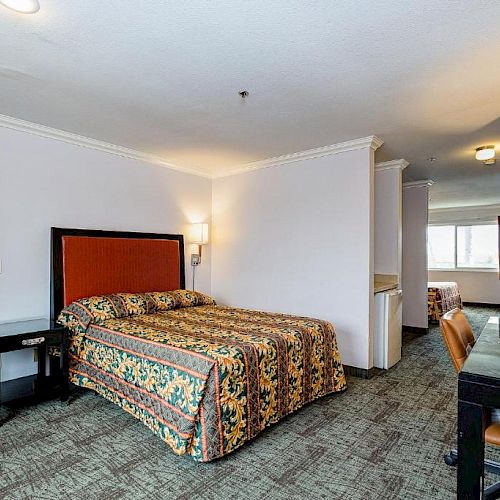 A hotel room with a bed, nightstands, a desk with a chair, a lamp, and a carpeted floor. A small kitchenette area is visible in the background.