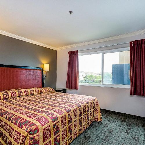 A hotel room with a king-sized bed, patterned bedspread, wall lamps, window with curtains, and a dresser.