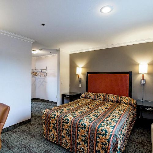 The image shows a well-lit hotel room with a patterned bedspread, desk and chair, bedside tables, a closet area, and a window with curtains.