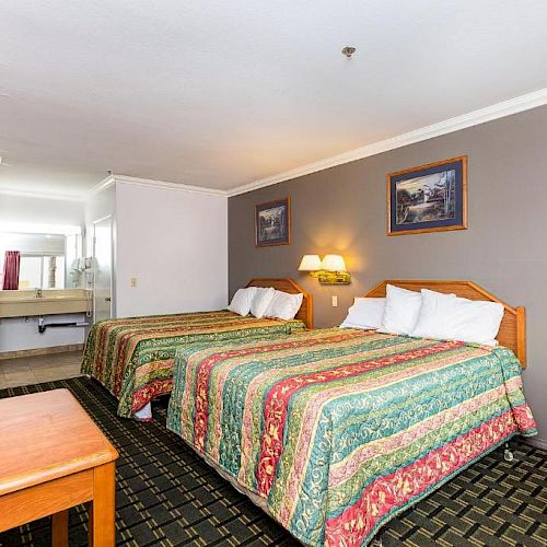 A hotel room with two beds, a TV on the wall, a wooden desk, and a sink and mirror area in the background.