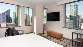 A modern bedroom with large windows, a city view, a bed, a chair, a wall-mounted TV, and minimal furniture completes the scene.