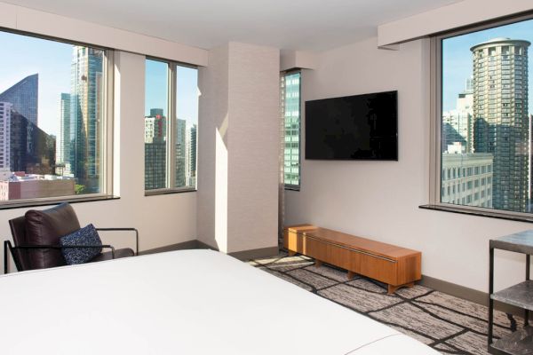 A hotel room with a large bed, TV, chair, and city view.