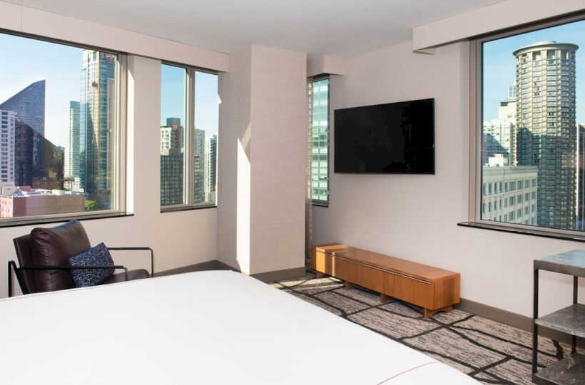 A modern bedroom with large windows, a city view, a bed, a chair, a wall-mounted TV, and minimal furniture completes the scene.