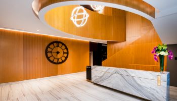 A modern lobby with a marble reception desk, wooden accents, a large wall clock, contemporary lighting, and a flower arrangement.