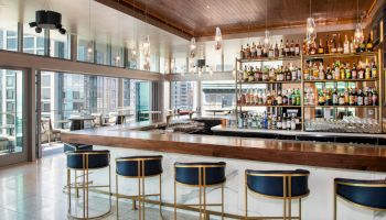 The image shows a modern bar with a wooden counter, high chairs, and shelves stocked with bottles behind the counter, overlooking tall buildings outside.