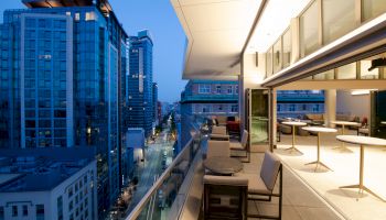 A modern cityscape at dusk with tall buildings, viewed from a spacious balcony with outdoor seating and tables.
