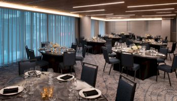 The image shows an elegantly set banquet hall with round tables, black chairs, white table settings, and floral centerpieces under modern lighting.