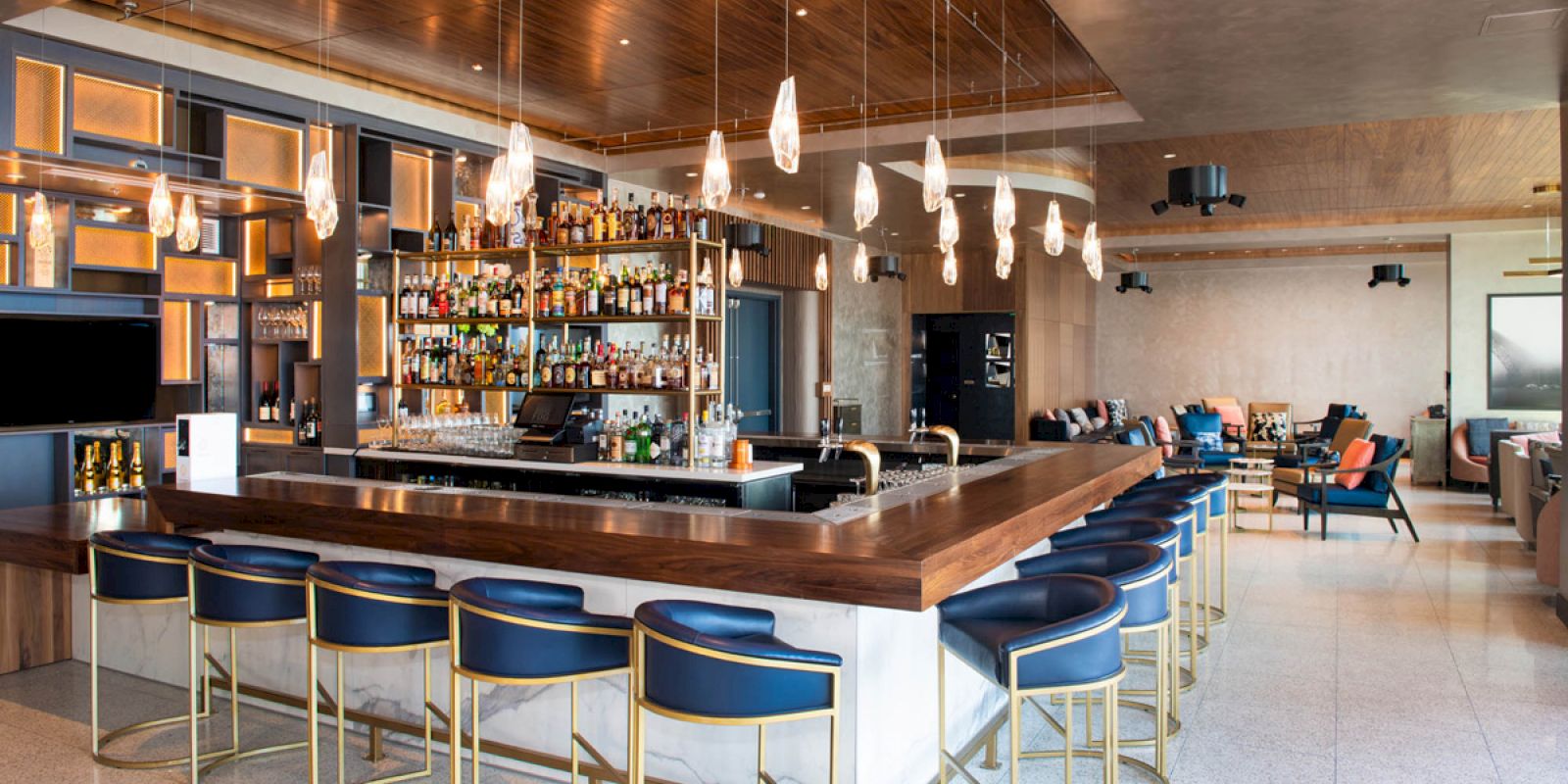 A modern bar with a wooden counter, blue bar stools, a variety of bottles, pendant lights, and a seating area with couches in the background.