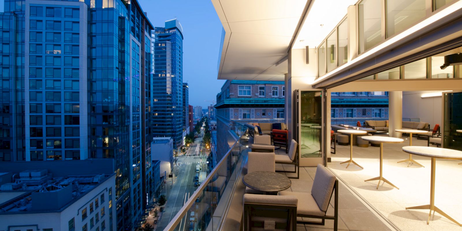 It's a cityscape view featuring modern buildings and a lit outdoor terrace with tables and chairs during the evening.
