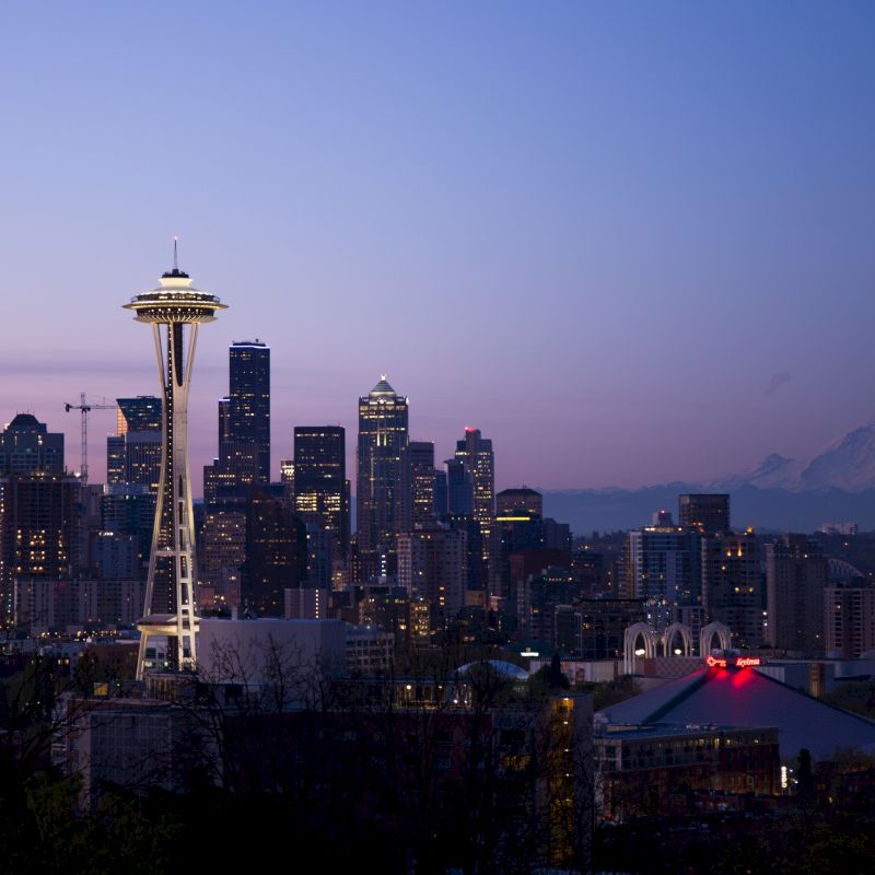 The image shows the skyline of Seattle at dusk with the Space Needle prominently visible, with Mount Rainier in the background under a clear sky.